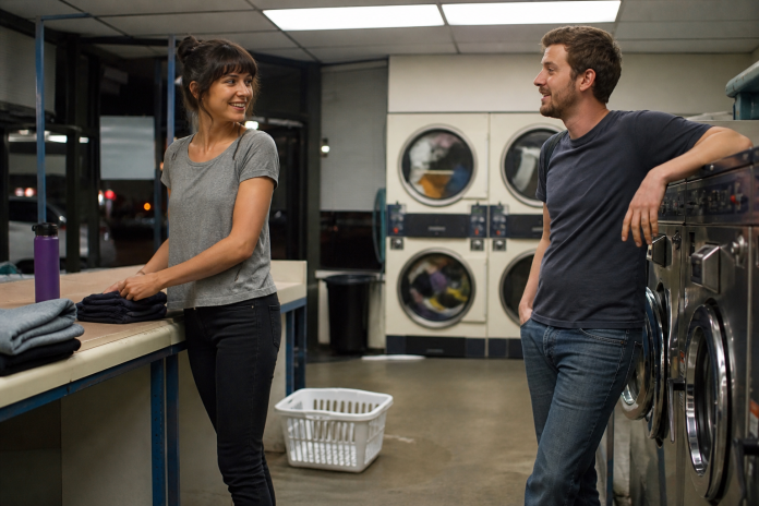 Two people in their thirties talking in a laundromat at night, one folding clothes and one leaning on a washing machine