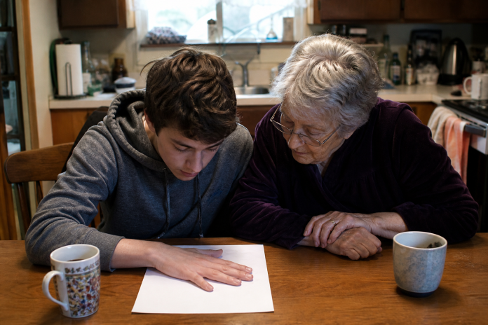 Grandmother and grandson trying the hand on white paper aura exercise Teenage boy and his grandmother at a kitchen table peering at his hand resting on white paper, tea mugs beside them