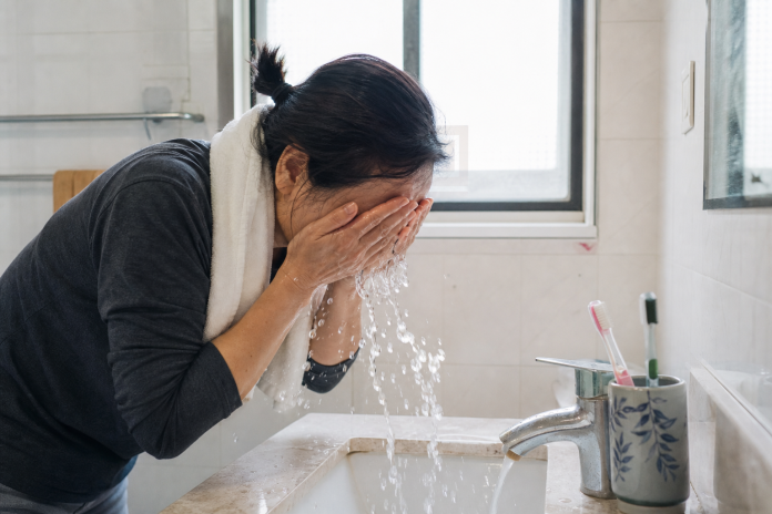 Woman in her forties splashing cold water on her face at a bright bathroom sink in the morning, towel over her shoulder