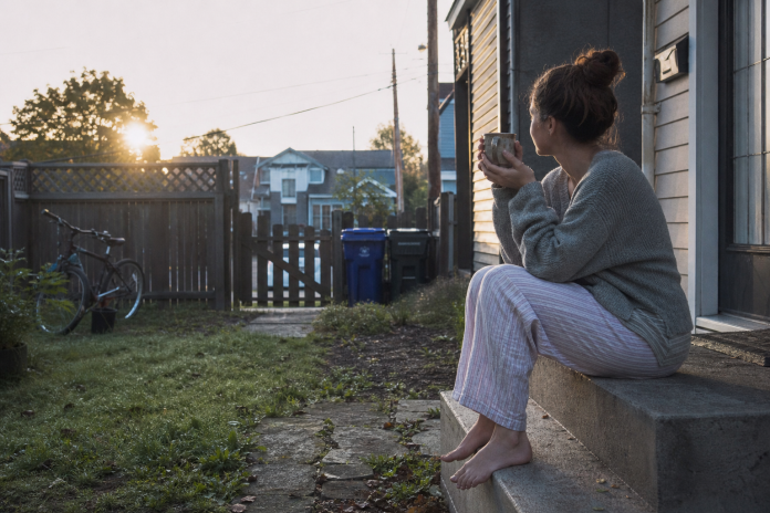 Woman in her late twenties sitting barefoot on a townhouse back step at sunrise with a coffee, bike and recycling bins in the small yard