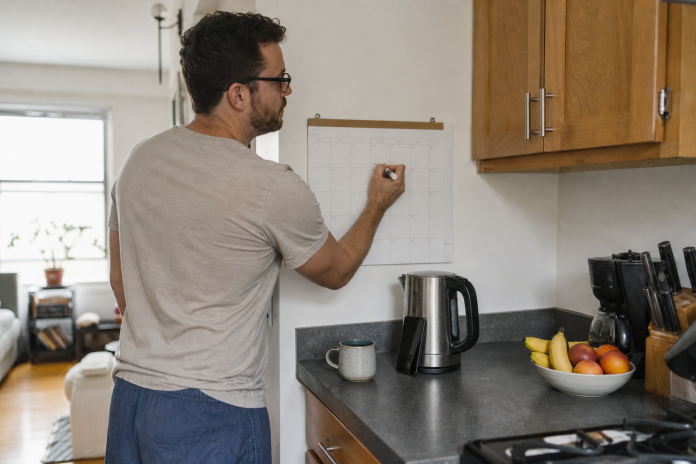 Manifesting your dreams: planning the week on a wall calendar Man in his thirties writing on a wall calendar at a small apartment kitchen counter in the morning, phone propped against the kettle