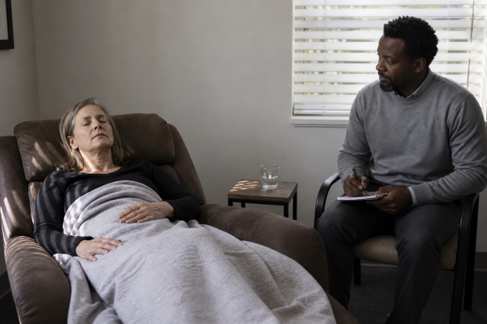 Woman in her fifties reclined with eyes closed under a blanket in a plain therapist's office while the practitioner sits to the side with a notepad, daylight through blinds