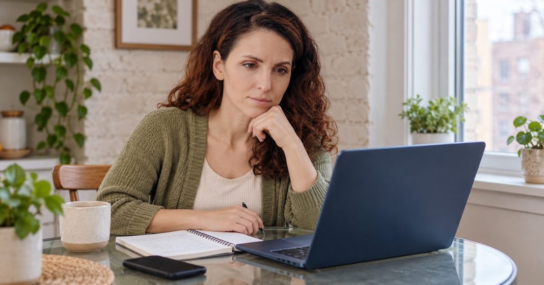 A woman comparing information on a blue laptop with an open notebook at a glass table at home.
